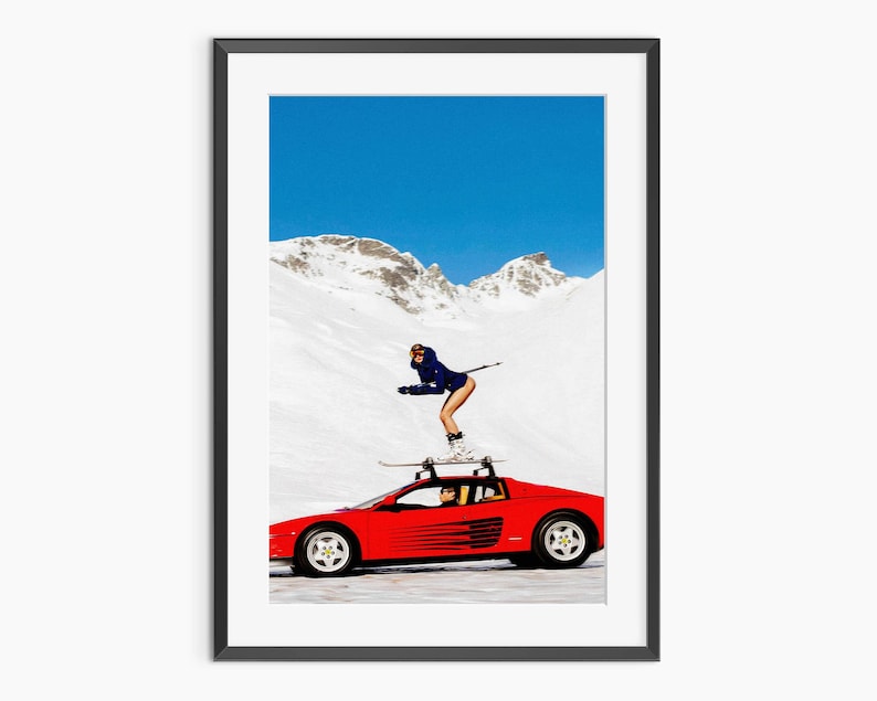A person is skiing on top of a red sports car in a snowy mountain landscape.