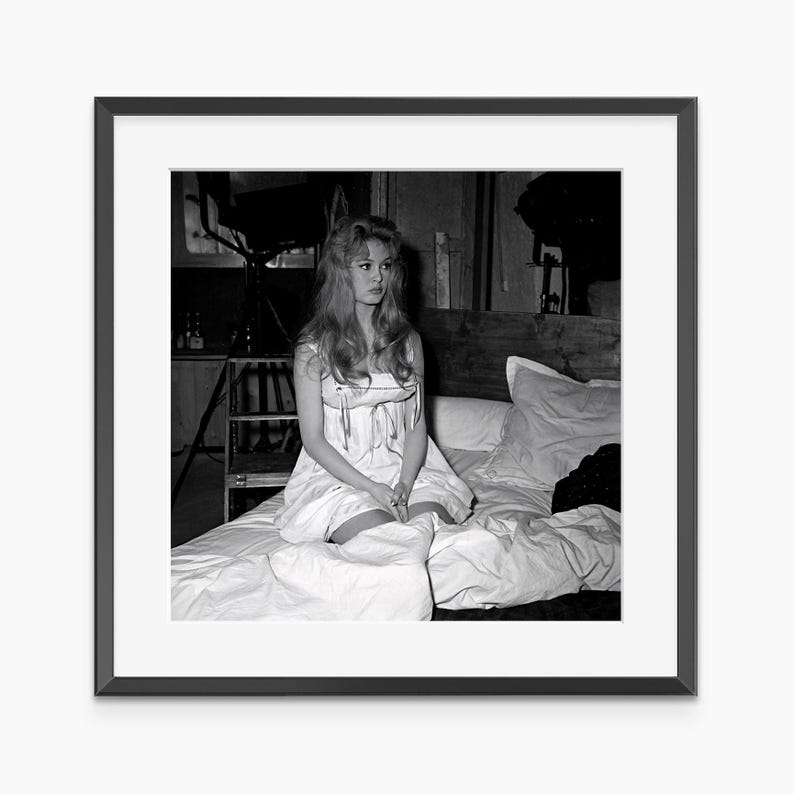 A woman in a white dress sits on a bed, her gaze directed off to the side.