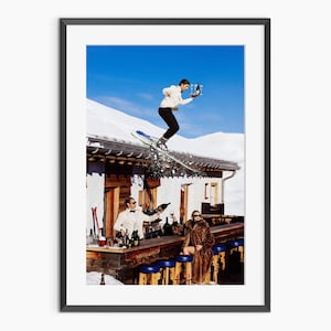May include: A framed print showing a winter scene with a person on skis jumping off a snowy roof, holding drinks. Below, a bar scene with people in formal attire. The sky is a clear blue.