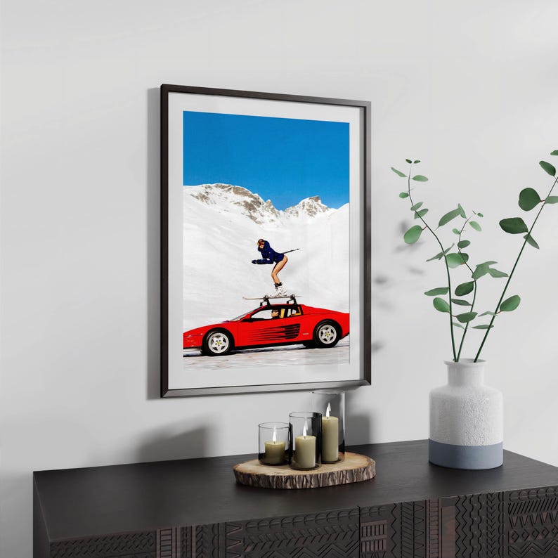 a framed poster of a woman skiing on a red sports car, with a mountainous background and a vase of green plants on a wooden surface below the frame.