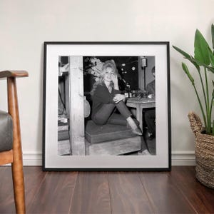 A framed black and white photograph of a woman sitting on a bench, with a potted plant and a wooden chair visible in the background.