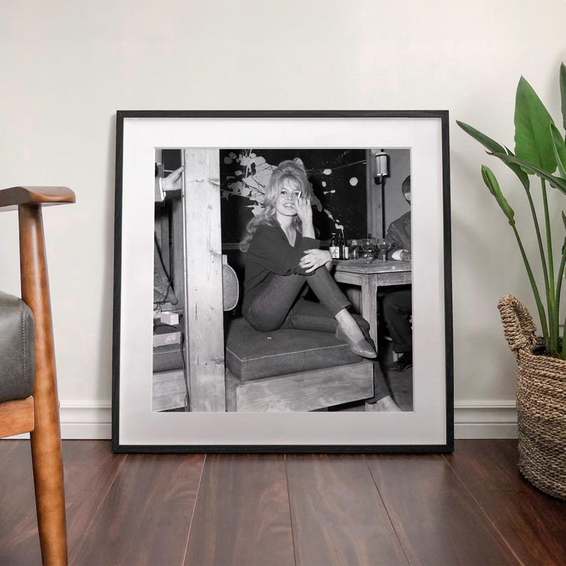 A framed black and white photograph of a woman sitting on a bench, with a potted plant and a wooden chair visible in the background.