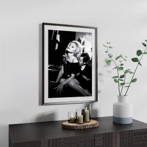 a framed black and white photograph of a woman sitting on a chair, with a potted plant and candles on a wooden surface below the frame.