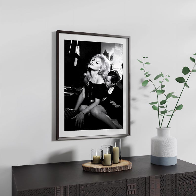 a framed black and white photograph of a woman sitting on a chair, with a potted plant and candles on a wooden surface below the frame.