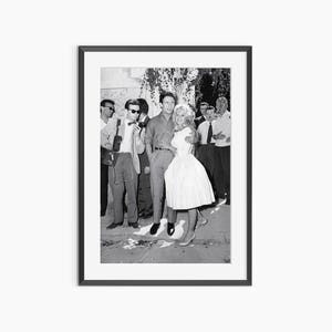 May include: Framed black and white photograph featuring a man and woman embracing, surrounded by a crowd. The woman is in a white dress, and the man wears a shirt. Other people in suits and sunglasses are in the background.