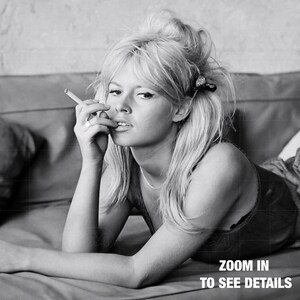 A black and white image of a woman lying on a couch, smoking a cigarette and looking directly at the camera.