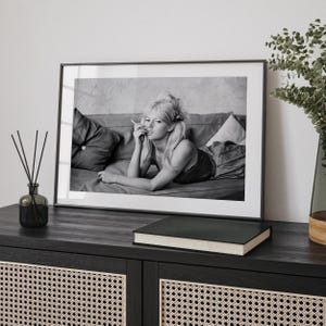 a framed black and white photograph of a woman lying on a couch, with a book and a vase on a shelf in the foreground.