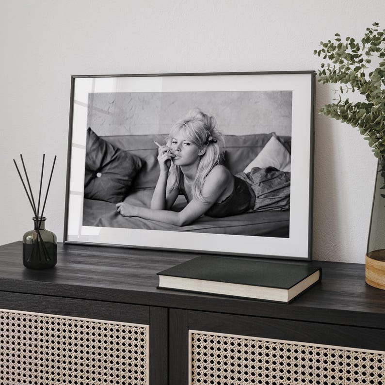 a framed black and white photograph of a woman lying on a couch, with a book and a vase on a shelf in the foreground.