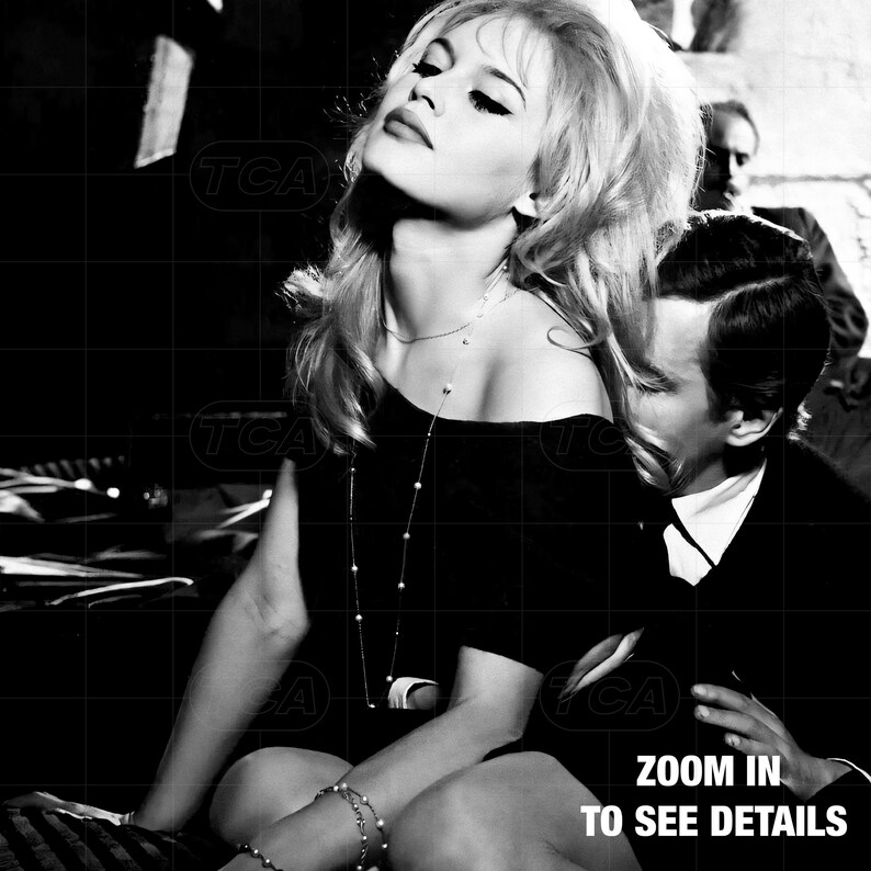 A black and white image of a woman with long blonde hair wearing a black dress, sitting next to a man in a suit.