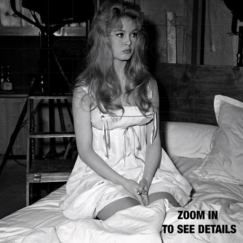 A black and white photograph of a woman sitting on a bed, wearing a white dress with a bow detail.