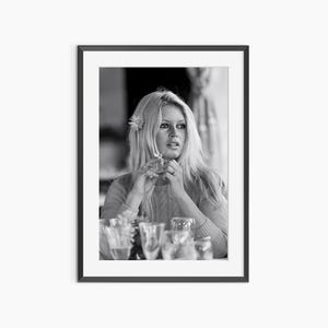 A black and white photograph of a woman sitting at a table, with wine glasses and a flower in her hair.