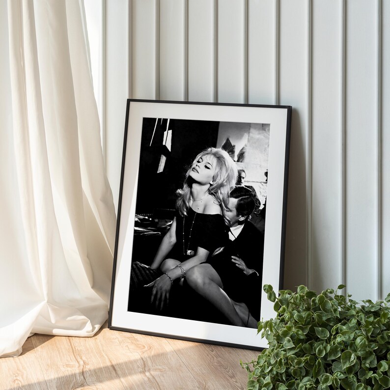 A framed black and white photograph of a woman sitting on a chair, with a man sitting next to her, is displayed on a wooden floor next to a plant.