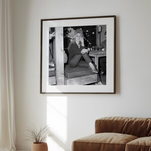 a framed black and white photograph of a woman sitting on a bench, with a potted plant and a couch visible in the foreground.