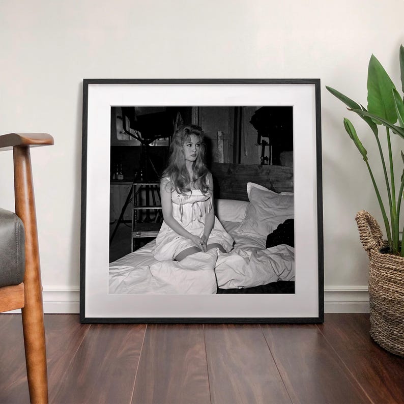 A framed black and white photograph of a woman sitting on a bed, with a potted plant to the right of the frame.