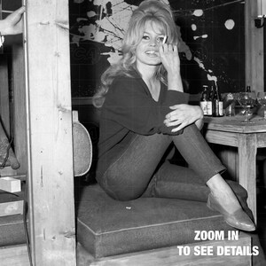 A black and white photograph of a woman sitting on a chair, with a mirror and various objects in the background, including bottles and wine glasses.