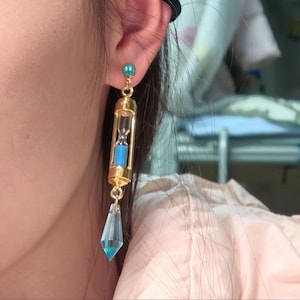 Könnte beinhalten: Goldfarbene Sanduhr-Ohrringe mit blauen Akzenten. Die Ohrringe haben einen kleinen blauen Edelstein oben, eine gläserne Sanduhr mit blauem Sand und einen hängenden blauen Kristall unten. Die Ohrringe sind zum Tragen am Ohr gedacht.