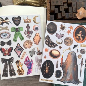 May include: Two sheets of decorative stickers with various gothic and celestial designs. Includes bows, butterflies, moons, eyes, ghosts, and portraits. The text "THE LOVERS" is visible on one sheet.