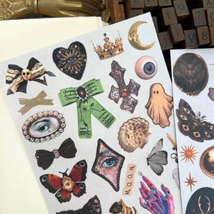 May include: A sheet of decorative stickers featuring various gothic and celestial designs. Includes a black heart with an eye, a gold crown, a crescent moon, a cross, a ghost, butterflies, and a green ribbon with text. The word "MOON" is spelled out with letter tiles.