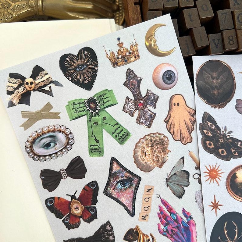 May include: A sheet of decorative stickers featuring various gothic and celestial designs. Includes a black heart with an eye, a gold crown, a crescent moon, a cross, a ghost, butterflies, and a green ribbon with text. The word "MOON" is spelled out with letter tiles.