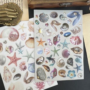 Puede incluir: Dos hojas de pegatinas decorativas con varias conchas marinas, estrellas de mar y perlas en tonos rosa, azul y dorado. Las pegatinas tienen un acabado brillante y están dispuestas sobre un fondo blanco. La imagen incluye una caja de madera y un objeto de latón de estilo vintage.