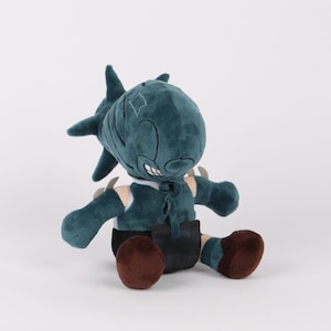 Puede incluir: Un peluche turquesa en posición sentada, con una cabeza con forma de tiburón y aletas puntiagudas. Viste un chaleco verde oscuro, pantalones cortos negros y tiene pies redondeados marrones. El juguete está sobre un fondo blanco liso.