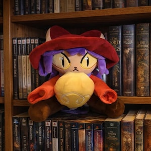 May include: A plush toy of a character with a red hat, orange sleeves, purple hair, and yellow eyes, holding a yellow sphere. The toy is set against a backdrop of a wooden bookshelf filled with books and video game cases.