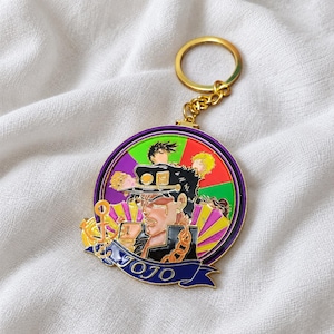 Puede incluir: Un llavero circular de metal con un diseño de esmalte colorido de un personaje con gorra y abrigo oscuros. El fondo incluye segmentos de color morado, verde, rojo y amarillo. Un icono de ancla dorada está a la izquierda y una banda azul dice JOJO.