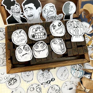 Puede incluir: Una colección de pegatinas de memes en blanco y negro con diversas expresiones faciales. Las pegatinas están dispuestas en una caja de madera y sobre sellos de letras de madera. Las expresiones van desde sonriendo hasta llorando.