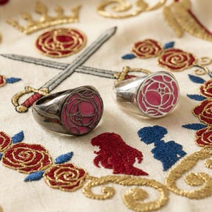 Könnte beinhalten: Zwei Ringe mit einem rosa Rosenmuster, einer schwarz und einer silberfarben, liegen auf einem cremefarbenen Stoff mit gestickten Rosen, Schwertern und Kronen. Die Ringe haben eine runde Oberfläche mit einem rosa Rosenmuster.