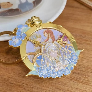May include: A gold-toned metal keyring featuring a circular clock face design with Roman numerals. The centre displays an anime-style illustration of three figures in soft pastel colours, surrounded by clusters of blue flowers and gold clock hands.