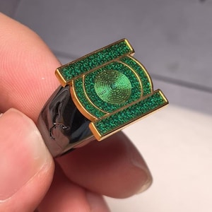 May include: A ring featuring a gold-toned rectangular face with a central circular emblem, all filled with a textured, shimmering green inlay. The band of the ring is a smooth, dark metallic finish, providing a contrast to the bright green face.