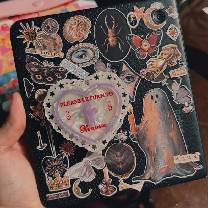 May include: Black tablet case decorated with various stickers, including a ghost holding a candle, butterflies, moons, and the text "PLEASE RETURN TO Heaven". The case has a heart-shaped embellishment and other celestial-themed stickers.