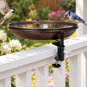 May include: A round, copper-colored bird bath basin filled with water, featuring a small decorative bird figurine on the rim. The basin is attached to a black metal clamp mount with a tightening screw, designed for securing to a deck railing.