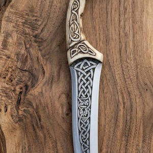 May include: A handcrafted knife with a bone handle and a stainless steel blade. The handle features intricate Celtic knotwork carvings. The blade has a curved shape and is set against a wooden background.