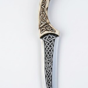 May include: A decorative knife with a curved handle and a silver blade. The handle is a light tan color with intricate Celtic knot designs carved into it. The blade also features Celtic knot patterns.