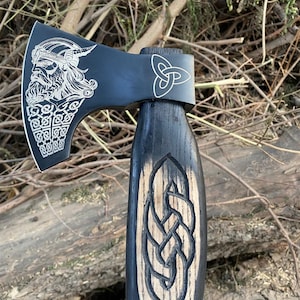 May include: A black axe with a wooden handle. The axe head features a detailed engraving of a Viking warrior and Celtic knot designs. The handle has a carved Celtic knot design. The axe is displayed outdoors.