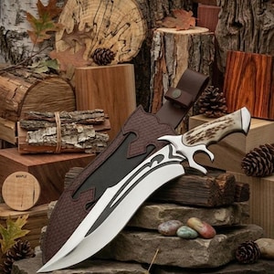 Könnte beinhalten: Ein dekoratives Messer mit gebogener Klinge und Griff mit hellbraunem und weißem Muster, in einer braunen Lederscheide. Das Messer wird mit Holzblöcken, Blättern und kleinen Steinen präsentiert.
