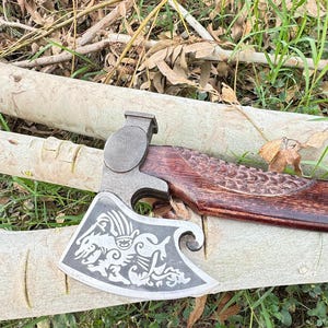 以下が含まれることがあります： 暗い木製の柄と銀色の刃が付いた手作りの斧。刃には神話上の生き物の彫刻が施されています。斧は丸太の上に置かれており、背景には草や葉があります。