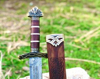 May include: A decorative sword with a patterned blade and a wooden handle. The handle is wrapped in brown leather with a decorative metal guard. The sword is displayed with its wooden sheath, which has an intricate carved design.