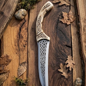 Könnte beinhalten: Ein handgefertigtes Messer mit einem Knochengriff und einer Edelstahlklinge. Der Griff weist aufwendige keltische Knotenmuster auf. Das Messer liegt auf einer Holzoberfläche mit Herbstblättern und Moos.