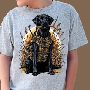 May include: A gray t-shirt featuring a black Labrador Retriever illustration. The dog is wearing a camouflage vest and holding antlers in its mouth. The background includes tall grass and a setting sun.