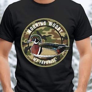 Może przedstawiać: Czarna koszulka z motywem kaczki karolinki. Projekt przedstawia ilustrację kaczki karolinki w okrągłym emblemacie. Tekst "MORNING WOODY MY FAVORITE" otacza kaczkę na tle kamuflażu. Kaczka ma kolorowe upierzenie.
