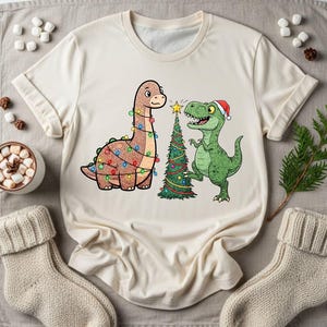 May include: Cream-colored t-shirt featuring a cartoon illustration of two dinosaurs and a Christmas tree. One dinosaur is brown with colorful lights, and the other is green with a Santa hat. A festive holiday tee.