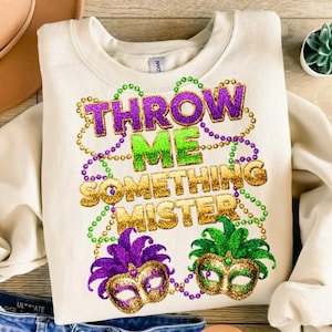 May include: A cream-coloured sweatshirt featuring the text "THROW ME SOMETHING MISTER" in glittery purple, green, and gold. The design includes Mardi Gras masks and bead strands. A festive apparel item.