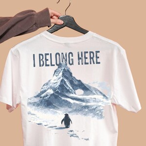 Könnte beinhalten: Weißes T-Shirt mit einer Berg- und Pinguin-Grafik. Der Text "I BELONG HERE" ist über dem Berg aufgedruckt. Der Pinguin geht im Schnee. Das T-Shirt hängt an einem schwarzen Kleiderbügel.