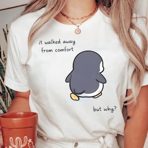 Könnte beinhalten: Weißes T-Shirt mit einem Cartoon-Pinguin-Design. Der Pinguin hat einen grauen Körper, gelbe Füße und einen weißen Bauch. Der Text "it walked away from comfort but why?" ist auf dem T-Shirt aufgedruckt. Ein lässiges Kleidungsstück.