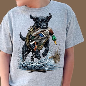 Op de afbeelding: Grijze t-shirt met een afbeelding van een zwarte Labrador Retriever die een eend draagt. De hond staat in het water, met spatten en riet. De eend heeft groene, blauwe en bruine veren.