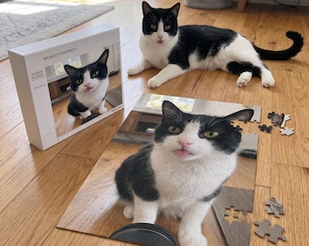 Peut inclure: Un puzzle représentant un chat noir et blanc avec la langue tirée, présenté à côté de sa boîte blanche marquée JIGSAW PUZZLES et HIGH QUALITY WOOD PRODUCTS, avec le vrai chat se reposant à proximité sur le plancher en bois.
