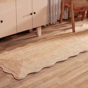 May include: A natural jute runner rug with a scalloped edge. The rug is rectangular with a light brown color and is placed on a wooden floor. The rug is near a white cabinet and a wooden chair.