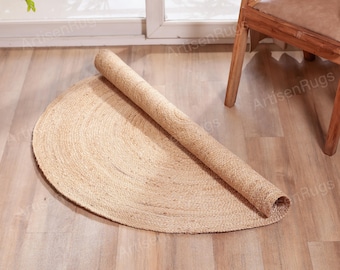 Puede incluir: Una alfombra de yute semicircular enrollada, en tonos beige natural, descansa sobre un suelo de madera clara. La textura de la alfombra es visible, y una silla de madera es parcialmente visible en el fondo. La alfombra es un revestimiento decorativo para el suelo.